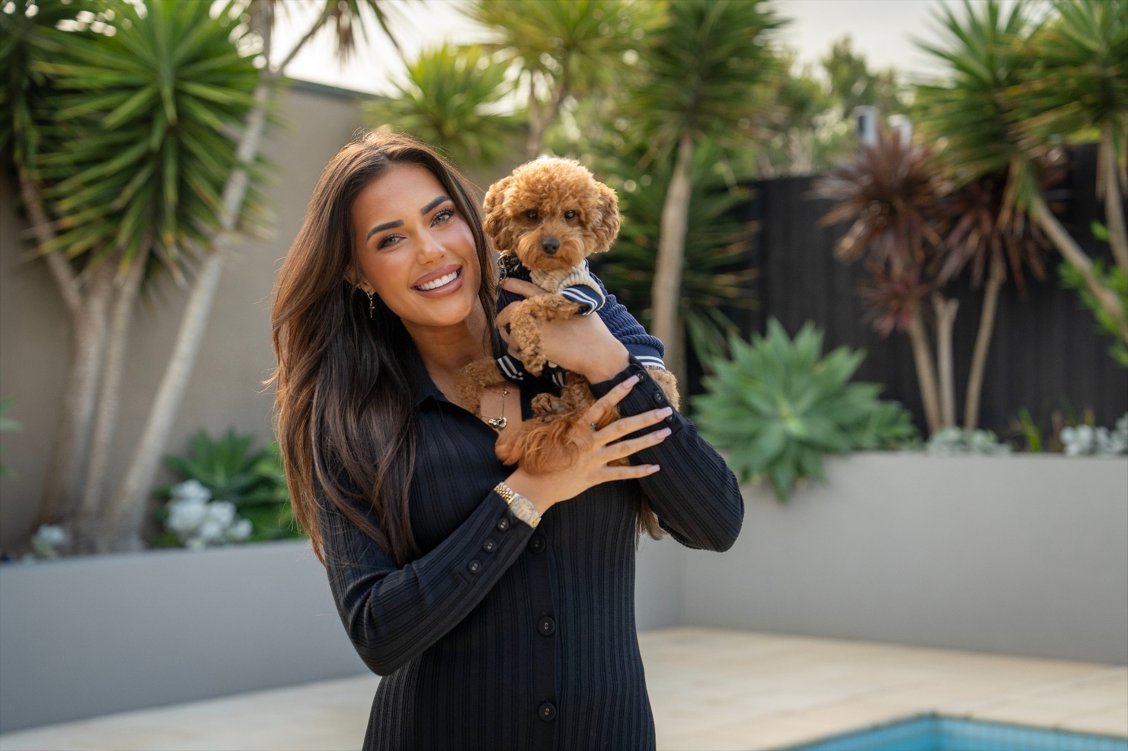 Woman holding a small dog in a garden setting with plants and a pool in the background