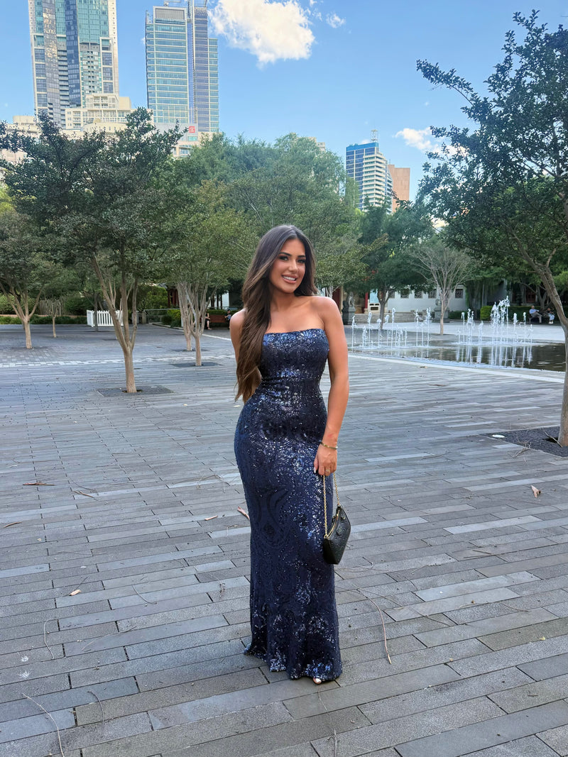 Woman in a blue sequin dress standing in an urban park with trees and buildings in the background.