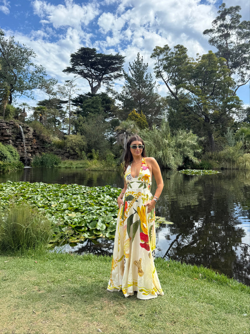 Claire Nomarhas is wearing a yellow and white tropical floral print halter maxi dress, standing on a grassy lawn next to a pond with lily pads and trees under a blue sky with white clouds.