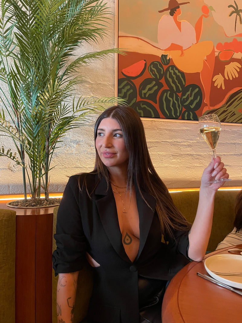 Claire Nomarhas is wearing a black open-front blazer and a gold necklace, holding a wine glass in a restaurant setting with a colorful mural on the wall.