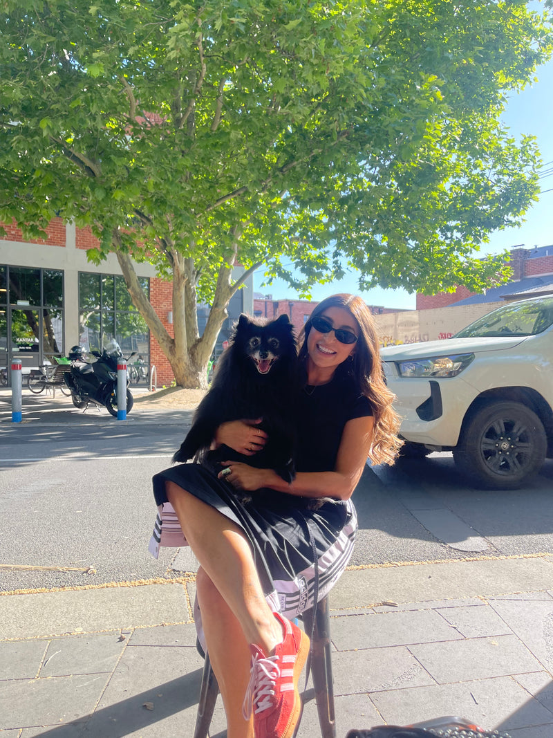 Claire Nomarhas is wearing a black sleeveless top, a black and white patterned skirt, and red sneakers is crouching outdoors on a sidewalk next to a street, holding a small black dog.