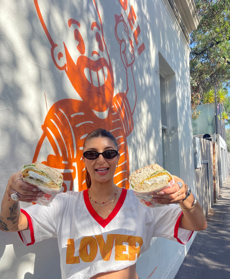 Claire Nomarhas is wearing a white t-shirt with red trim and the word 'LOVES' in orange, holding two large sandwiches outdoors in front of a white wall with an orange mascot graphic.
