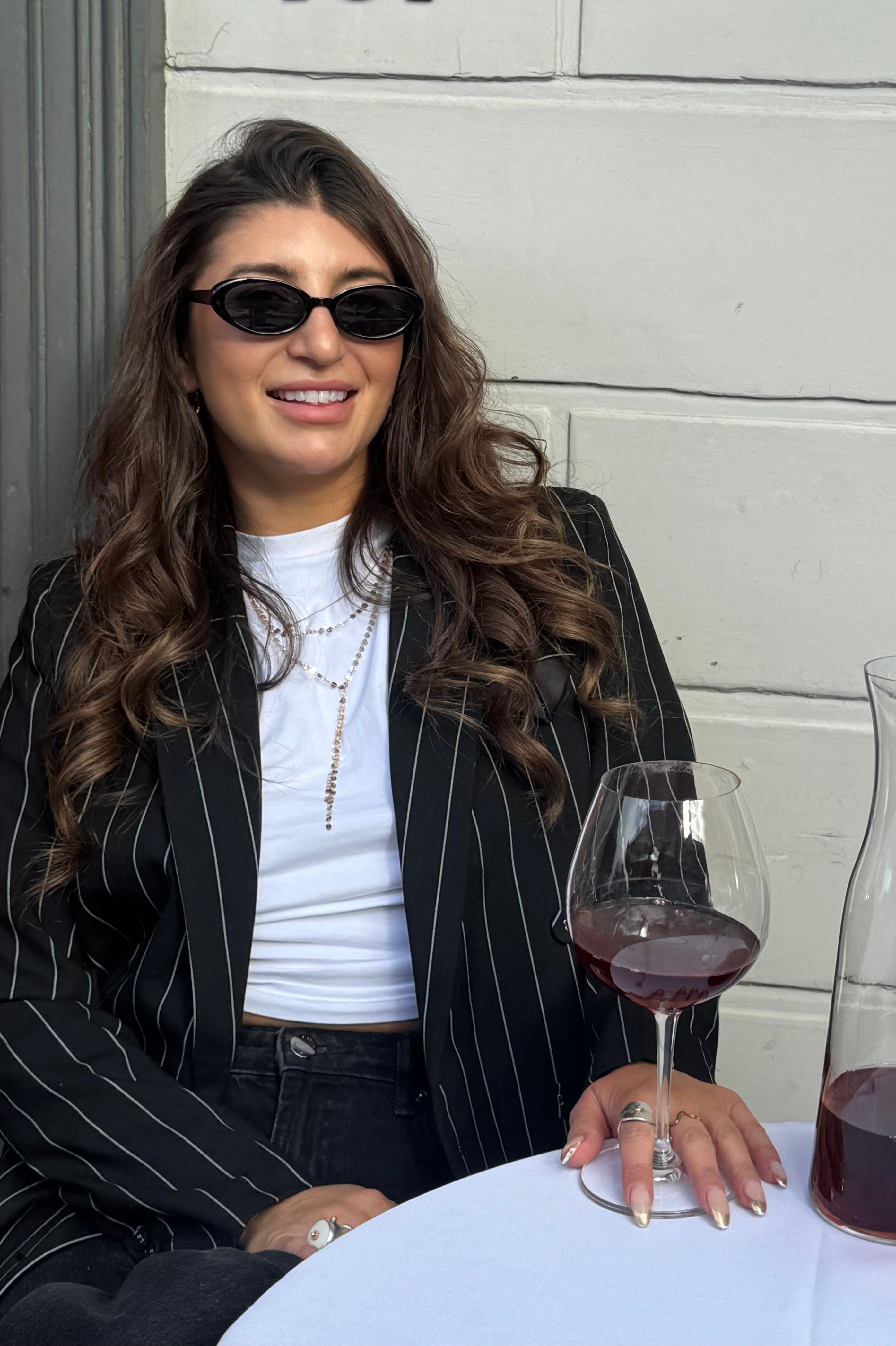 Claire Nomarhas is wearing black sunglasses, a white ribbed top, and a black pinstripe blazer sits at an outdoor table with wine glasses and a carafe against a white paneled wall with the number 181.