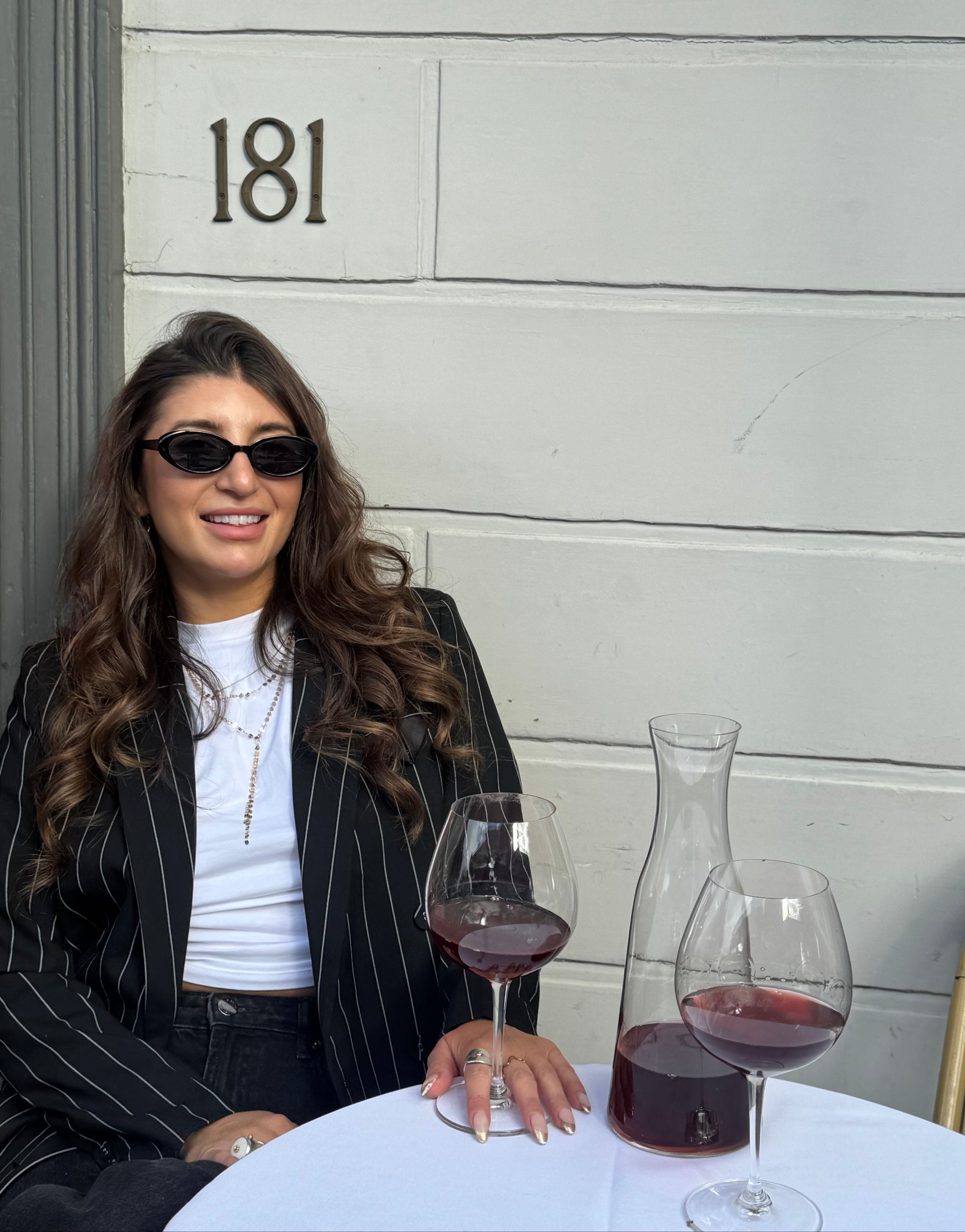 Claire Nomarhas is wearing black sunglasses, a white ribbed top, and a black pinstripe blazer sits at an outdoor table with wine glasses and a carafe against a white paneled wall with the number 181.