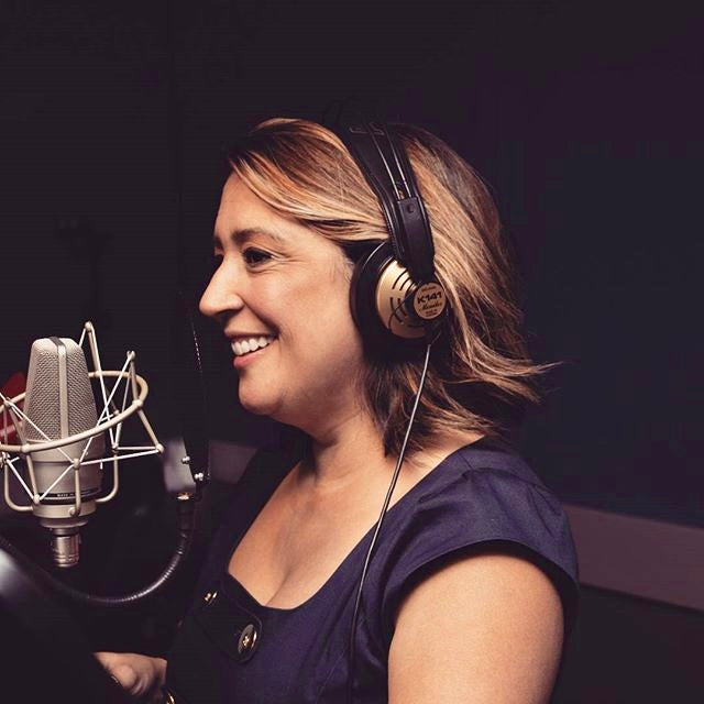Woman wearing headphones and smiling in front of a microphone in a recording studio.