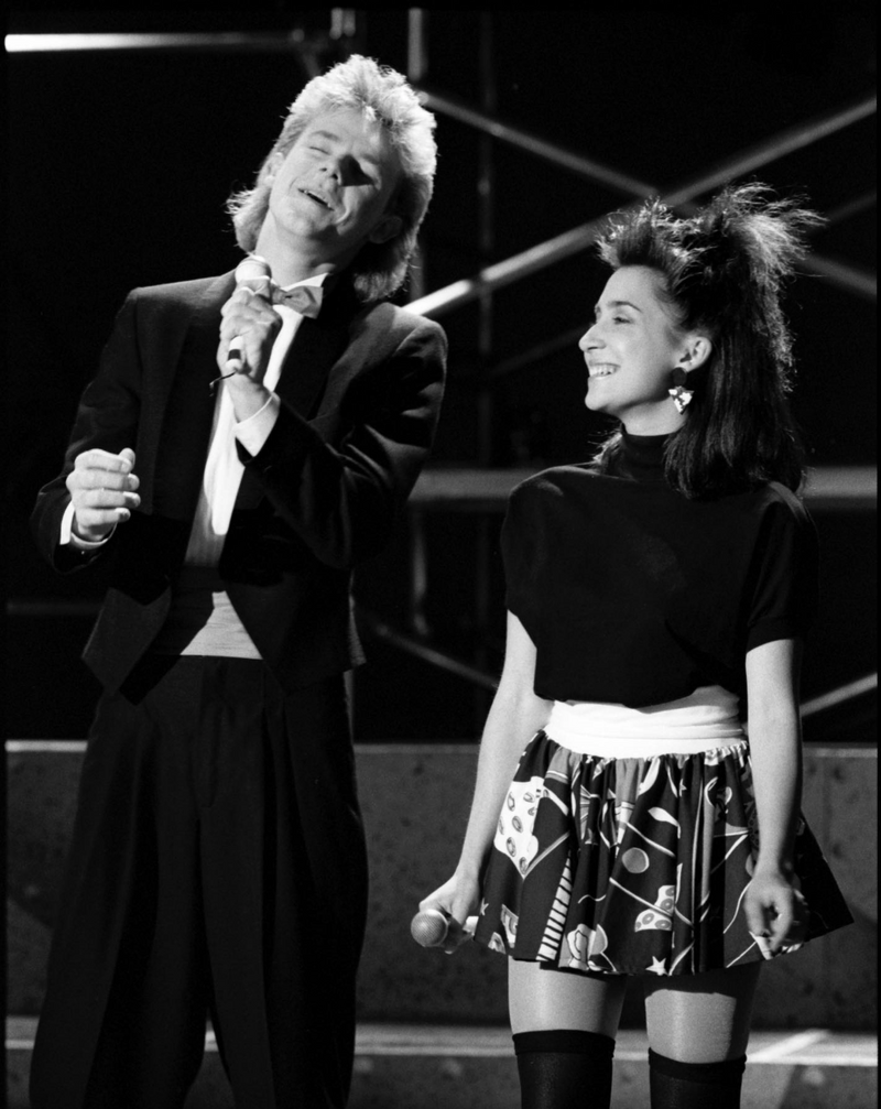 Two people on stage, one in a suit and the other in a patterned dress, with a black and white color scheme.