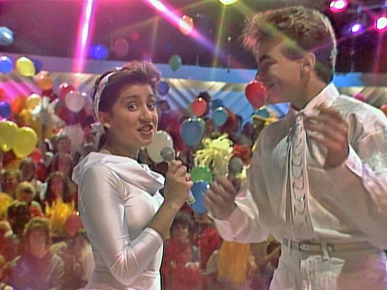 Two people in white outfits with a microphone and balloons in a festive setting