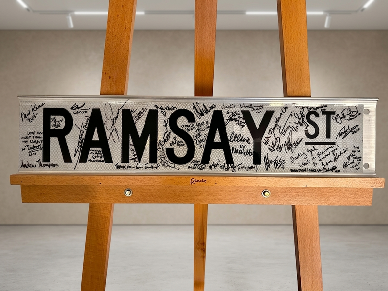 Street sign with 'RAMSAY ST' on a wooden stand against a neutral background