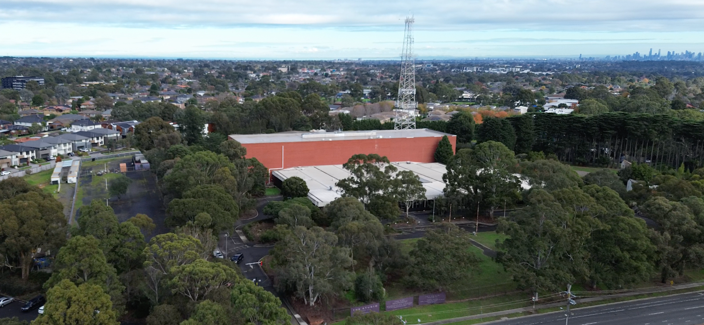 An aerial view of a large brick building with a white roof, surrounded by dense green trees and grass, with a city skyline visible in the distance under a cloudy sky.