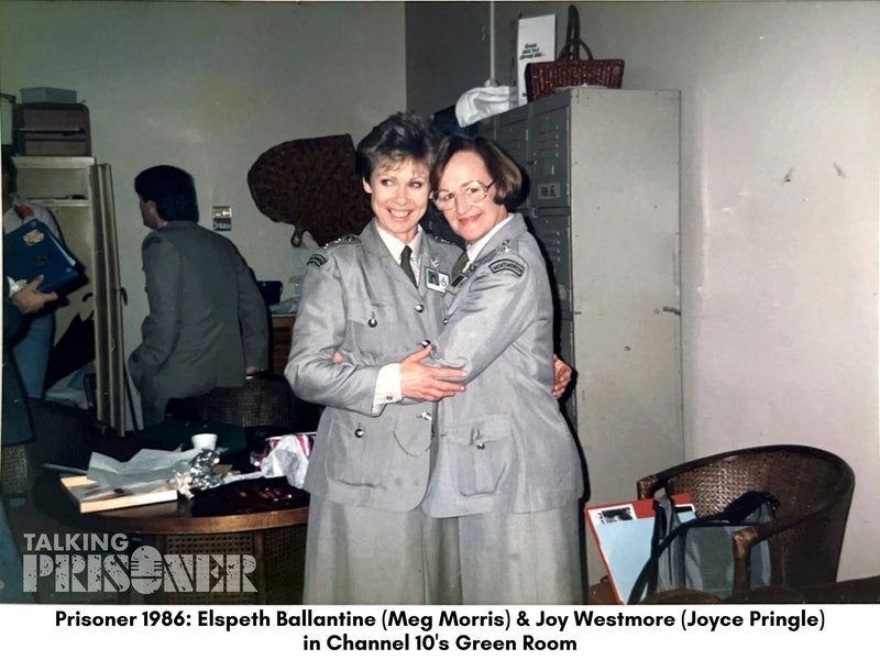 Two women in military uniforms posing together in a room with other people and furniture.
