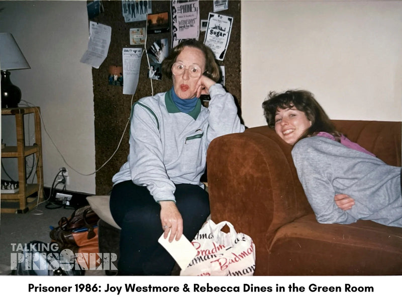 Two women sitting on a couch in a room with posters and a lamp.