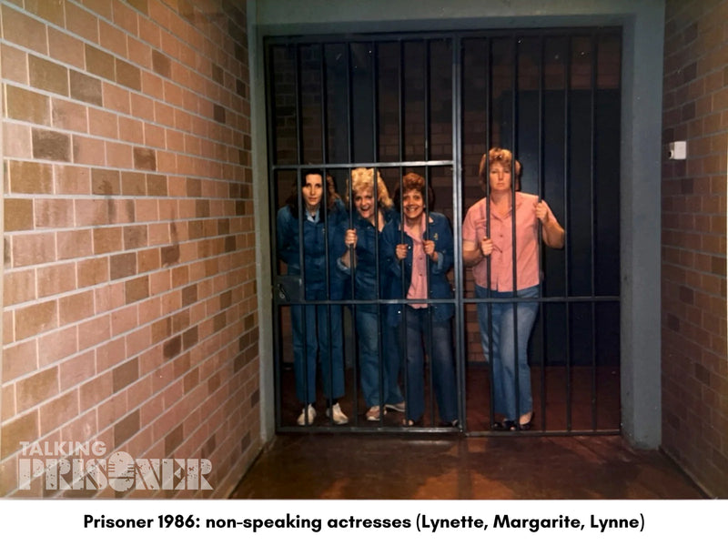 Four women in prison uniforms standing behind a barred door in a hallway with brick walls.