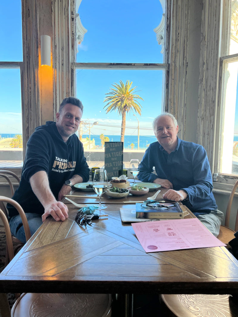 Two men sitting at a table with a scenic view of a palm tree and ocean through large windows.