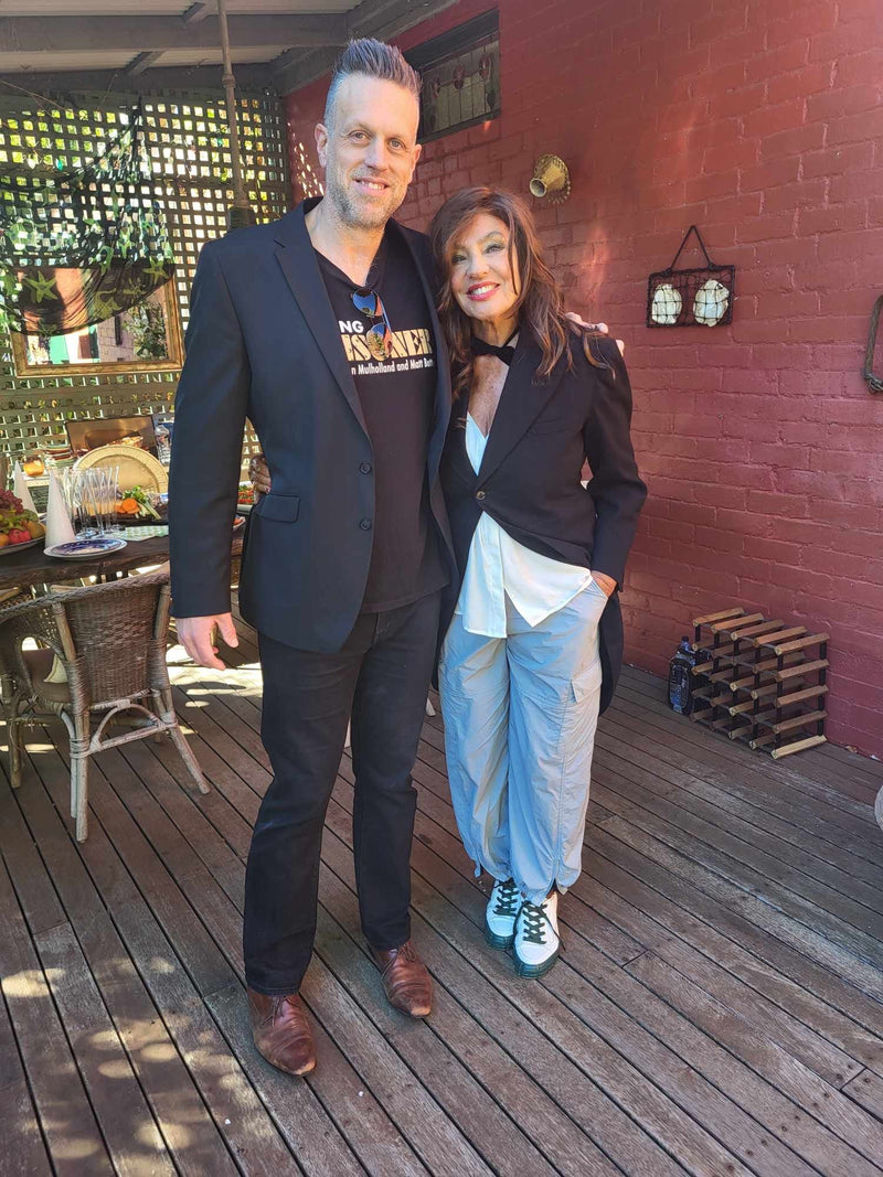 Two people standing on a wooden deck with a brick wall and outdoor furniture in the background.