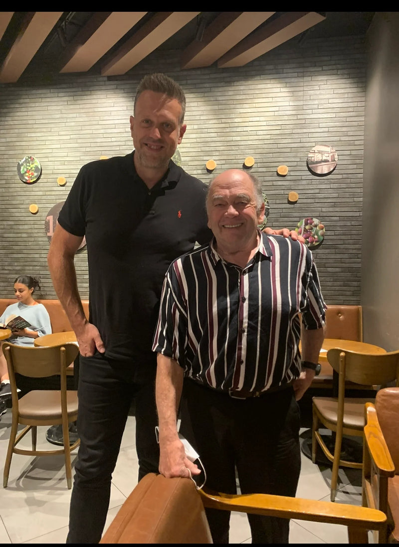 Two men standing together in a restaurant setting with wooden chairs and tables.