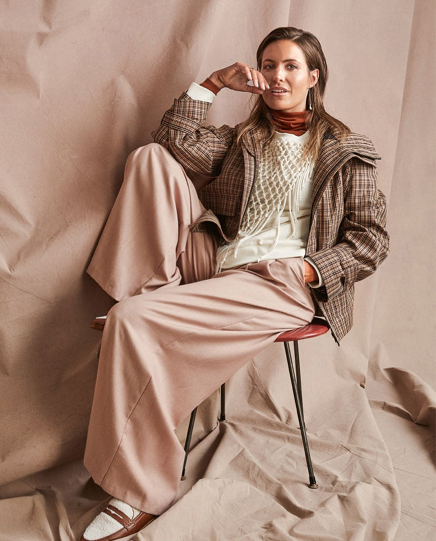A woman sits on a stool against a draped beige backdrop, wearing a brown plaid jacket, beige wide-leg trousers, a cream knit top, and white loafers with brown trim.
