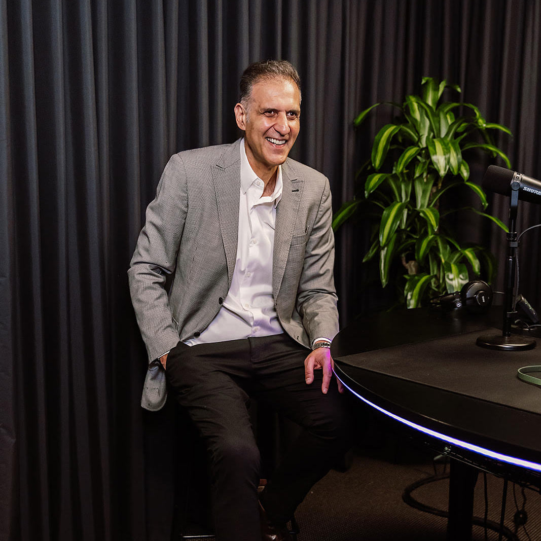 Man in a gray suit sitting on a chair in a studio setting with a plant and equipment in the background.