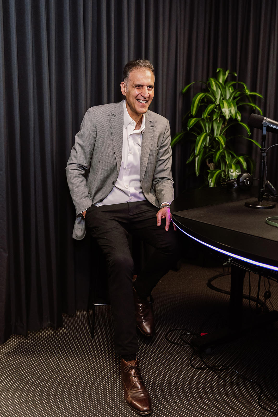 Man in a gray suit sitting on a chair in a studio setting with a plant and equipment in the background.